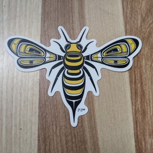 Puede incluir: Una abeja estilizada negra y amarilla con detalles intrincados, que se asemeja a un diseño nativo americano. La abeja está sobre un fondo blanco.
