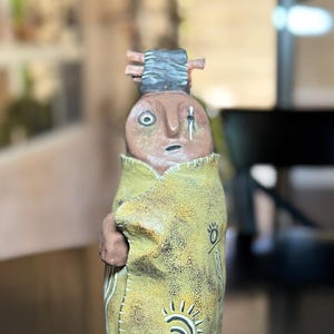 May include: A handcrafted ceramic figurine depicting a Native American figure. The figure has a brown face and is wrapped in a yellow cloak adorned with sun and bird motifs. The figurine is approximately 8 inches tall.