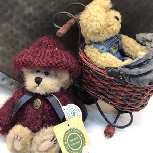May include: Two plush teddy bears, one wearing a burgundy knitted hat and jacket, the other wearing denim overalls, sitting in a wicker basket on a toy bicycle. The bear in the basket is holding a small blue bag. The bear in the hat has a tag that reads "Investment Collectibles The Archive Collection Kayla".