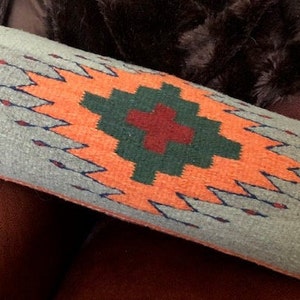 May include: A long, cylindrical pillow with a gray background and a colorful geometric pattern. The pattern features a large orange diamond with a green and red cross in the center.