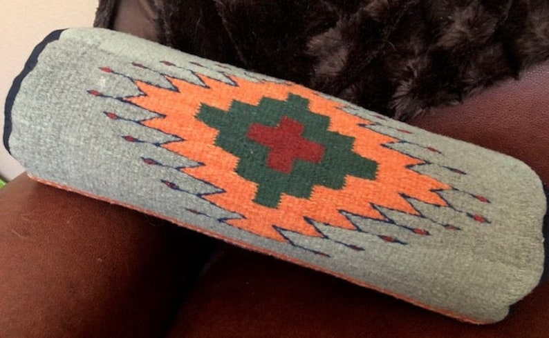 May include: A long, cylindrical pillow with a gray background and a colorful geometric pattern. The pattern features a large orange diamond with a green and red cross in the center.