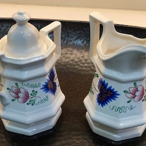 May include: A white ceramic creamer and sugar set with blue and pink floral designs. The creamer has a handle and a lid. The sugar bowl has a lid.