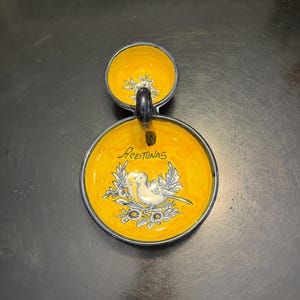 May include: A yellow ceramic serving dish with a small bowl attached. The larger bowl has a bird and floral design with the word "Aceitunas" in script. The dish has a dark blue handle and rim.