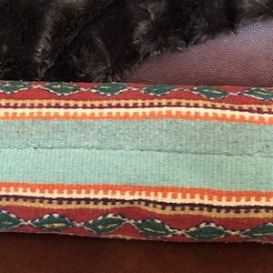 May include: A cylindrical pillow with a colorful woven pattern. The pillow has a red, orange, green, and blue striped pattern with a diamond pattern in the center. The pillow is made of a soft, textured fabric.
