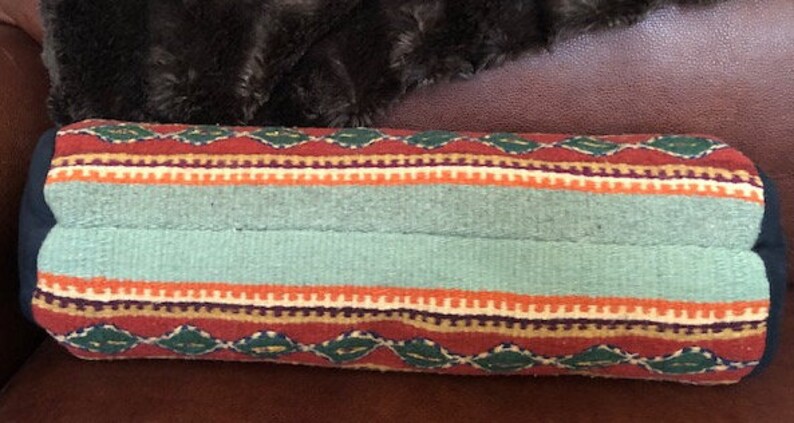 May include: A cylindrical pillow with a colorful woven pattern. The pillow has a red, orange, green, and blue striped pattern with a diamond pattern in the center. The pillow is made of a soft, textured fabric.