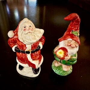 May include: A ceramic Santa Claus figurine with a red and white suit and a white beard, and a ceramic gnome figurine with a red hat, green pants, and a red apple in its hand.
