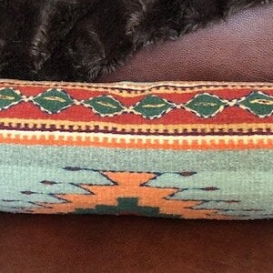 May include: A long, cylindrical pillow with a southwestern-style design. The pillow is made of woven fabric with a blue, orange, red, green, and white pattern.