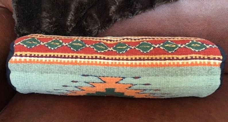 May include: A long, cylindrical pillow with a southwestern-style design. The pillow is made of woven fabric with a blue, orange, red, green, and white pattern.