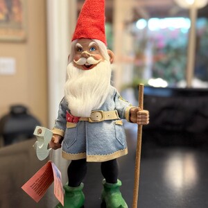 May include: A whimsical garden gnome figurine with a red pointed hat, white beard, and blue coat. The gnome holds a wooden staff and a decorative key. It wears green boots and a belt with a gold buckle.
