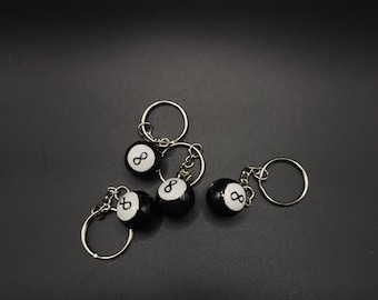 May include: A collection of five black and white 8-ball keychains. Each keychain features a small, round 8-ball charm with the number 8 in white. The keychains have silver-colored metal rings.