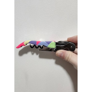 May include: A stainless steel corkscrew with a colorful, geometric patterned handle. The handle features pink, yellow, purple, green, and blue shapes. The corkscrew is held by a hand against a white background.