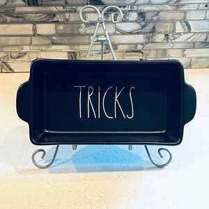 May include: Black ceramic baking dish with white lettering that says "TRICKS" on a silver stand.
