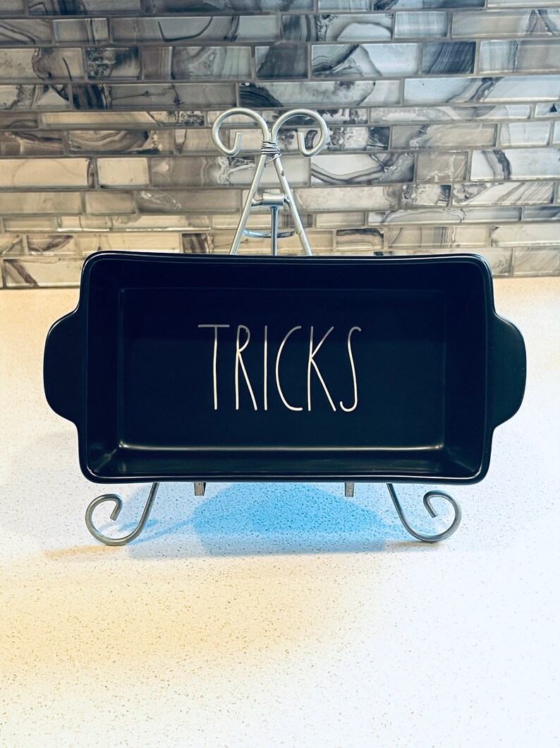 May include: Black ceramic baking dish with white lettering that says "TRICKS" on a silver stand.