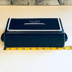 May include: A black rectangular baking dish with a white rim and a white handle on each side. The dish is 9 inches long. The dish has the text "RAE DUNN. ARTISAN COLLECTION" on the bottom.