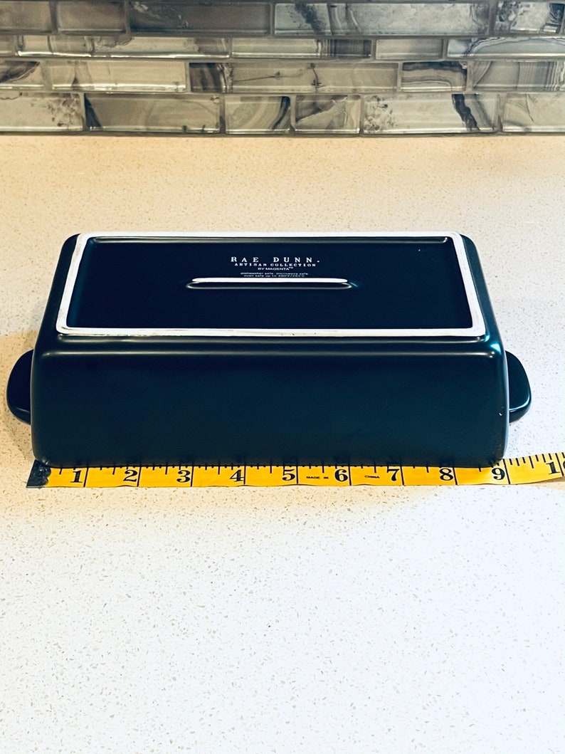 May include: A black rectangular baking dish with a white rim and a white handle on each side. The dish is 9 inches long. The dish has the text "RAE DUNN. ARTISAN COLLECTION" on the bottom.