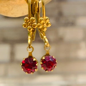 May include: A pair of gold dangle earrings with small red gemstone drops. The earrings have a flower-shaped design at the top.