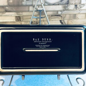 May include: Black rectangular baking dish with white trim. The dish is labeled "Rae Dunn. Artisan Collection by Magenta" and is dishwasher, microwave, and oven safe up to 400 degrees Fahrenheit.