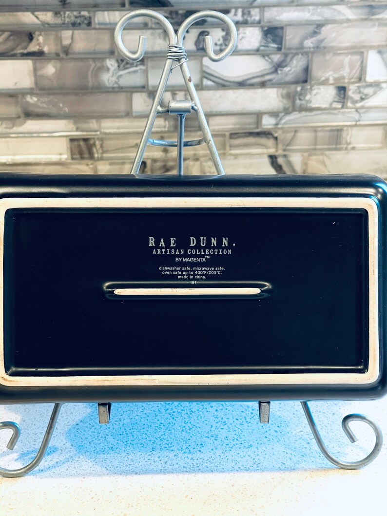 May include: Black rectangular baking dish with white trim. The dish is labeled "Rae Dunn. Artisan Collection by Magenta" and is dishwasher, microwave, and oven safe up to 400 degrees Fahrenheit.