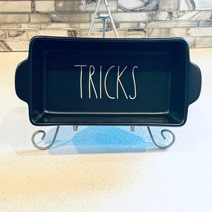 May include: Black ceramic baking dish with white lettering that says "TRICKS". The dish is sitting on a silver metal stand.