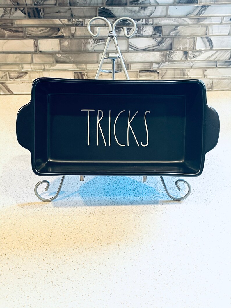 May include: Black ceramic baking dish with white lettering that says "TRICKS". The dish is sitting on a silver metal stand.