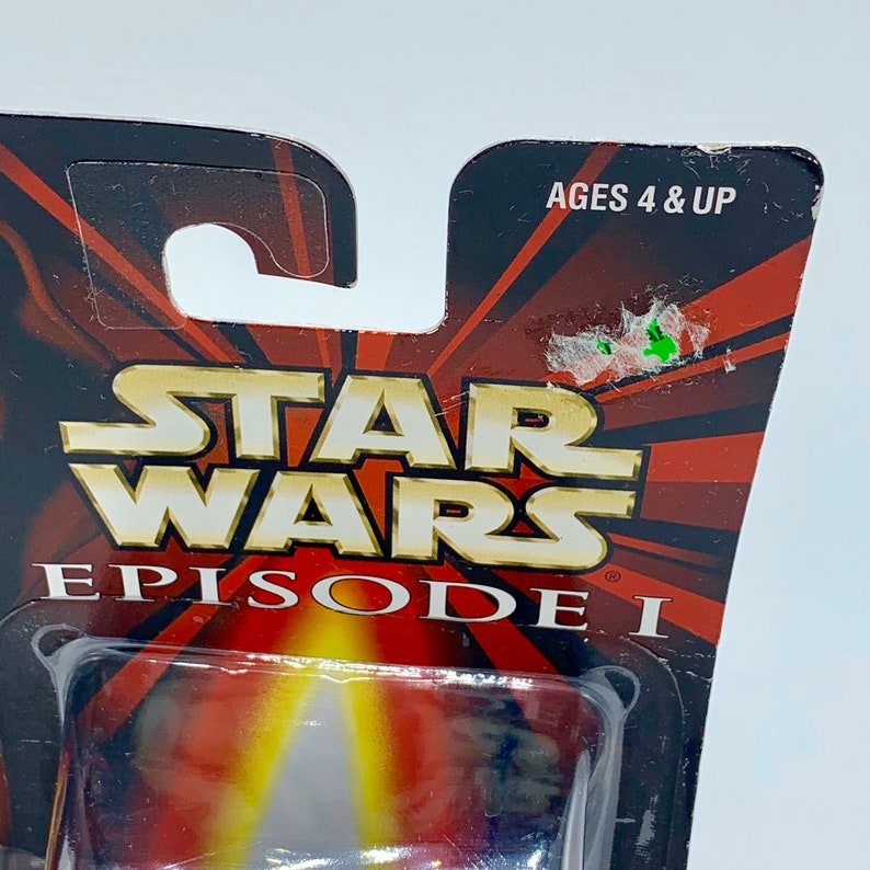 May include: A Star Wars Episode I toy packaging with a red, yellow, and black background. The packaging has a clear plastic window showing a toy lightsaber. The text on the packaging reads "Star Wars Episode I" and "Ages 4 & Up".