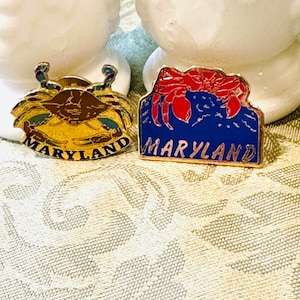 Puede incluir: Dos pines de recuerdo de Maryland en tono dorado con cangrejos azules. El pin de la izquierda tiene un cangrejo azul con pinzas marrones y un fondo azul y amarillo. El pin de la derecha tiene un cangrejo rojo con pinzas azules y un fondo azul. Ambos pines dicen "Maryland" en letras doradas.