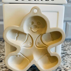 Pode incluir: Um cortador de biscoitos de gengibre de cerâmica bege. O cortador tem um design simples com uma cabeça redonda, dois braços e duas pernas.