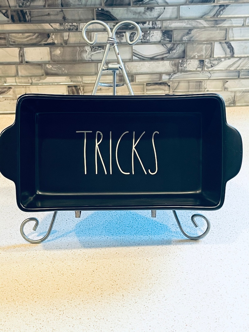May include: Black rectangular baking dish with white lettering that says "TRICKS" on a metal stand.