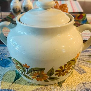 May include: A cream-colored ceramic sugar bowl with a matching lid and two side handles. The body features a speckled finish and a painted floral pattern around the base, consisting of orange and yellow daisy-like flowers with green leaves.