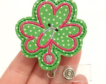 May include: Green shamrock with pink stitching and white polka dots, featuring a stethoscope design. The shamrock is attached to a clear plastic badge holder with a silver clip.