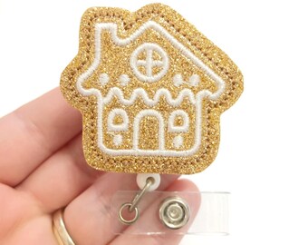 Puede incluir: Carrete de insignia de casa de jengibre con purpurina dorada. La casa de jengibre es blanca con purpurina dorada. El carrete de insignia es de plástico transparente con un clip de metal dorado.