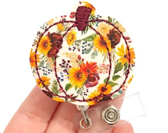 May include: A white pumpkin-shaped badge holder with a floral pattern featuring sunflowers, burgundy flowers, and greenery. The badge holder has a clear retractable badge reel.