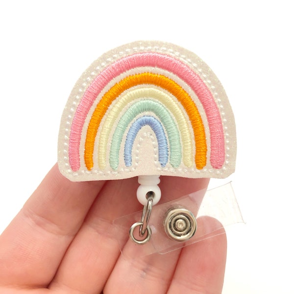 Könnte beinhalten: Ein weißer Regenbogen-Badge-Halter mit einem pastellfarbenen Regenbogen-Design. Der Regenbogen besteht aus rosa, orange, gelbem, grünem und blauem Garn. Der Badge-Halter hat ein transparentes Plastikband und einen Metallclip.