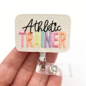 May include: White glitter badge holder with a rainbow embroidered "Athletic Trainer" text. The badge holder has a clear plastic retractable badge reel.