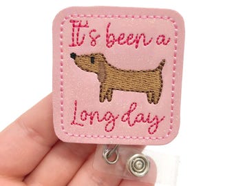 Puede incluir: Portagafete de purpurina rosa con un dachshund marrón y el texto "It's been a long day".