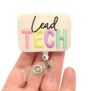 May include: A rectangular badge with the words "Lead TECH" embroidered in black, pink, yellow, purple, and green thread. The badge is cream-colored with a glittery texture and a white retractable reel with a clear plastic strap.