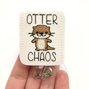 May include: White badge holder with a white background and black text that reads "Otter Chaos". A brown and white cartoon otter is embroidered in the center of the badge holder.
