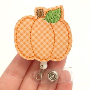 May include: Orange and white gingham pumpkin with a green leaf and brown stem, attached to a clear plastic badge holder with a silver clip.