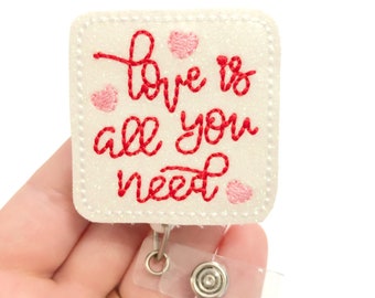 May include: White glitter badge holder with a red embroidered quote "Love is all you need" and pink hearts. The badge holder has a clear plastic retractable badge reel.
