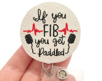 May include: White round badge with black and red embroidery. The text reads "If you FIB you get paddled" with a red EKG line and black paddles on either side. The badge is attached to a clear plastic retractable badge holder.