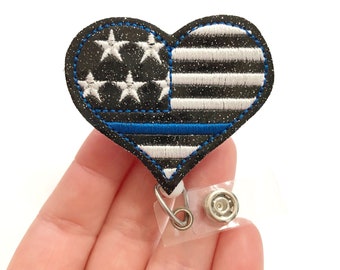 May include: A black and white glitter heart-shaped badge holder with a blue line and an American flag design. The badge holder has a clear plastic clip.