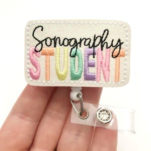 May include: A white glittery badge reel with the words "Sonography STUDENT" embroidered in black and pastel colors. The badge has a white border and is attached to a retractable reel with a clear plastic strap.