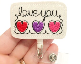 May include: White glitter badge reel with a white rectangle that says "love you" in black cursive with three embroidered hearts in red, purple, and pink.