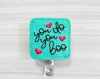 May include: A turquoise blue glitter badge holder with a black embroidered design that says "you do you boo" with three pink hearts. The badge holder is attached to a clear plastic retractable badge reel.