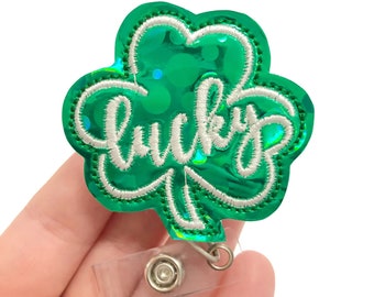 May include: Green iridescent shamrock with the word "lucky" embroidered in white. The shamrock is attached to a clear plastic badge holder with a metal clip.