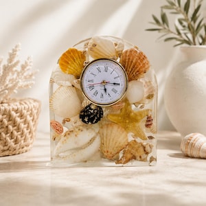 May include: A decorative clock encased in clear resin, featuring a variety of seashells in shades of white, tan, and orange. The clock face has Roman numerals and gold-colored trim. The clock is arched at the top, creating a unique coastal accent.