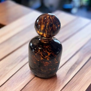 May include: A decorative glass bottle with a round stopper. The bottle is a dark amber color with a speckled pattern. The bottle has a rounded base and a cylindrical body.