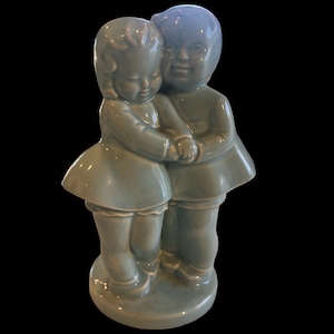 May include: A ceramic figurine of two children embracing, painted in a muted teal color. The figures are stylized with simple features and are standing on a round base. The children are holding hands and wearing dresses.