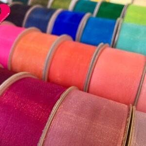 May include: A close-up of spools of colorful sheer ribbon with a glittery finish. The ribbon is arranged in a row, with the colors ranging from pink to purple.