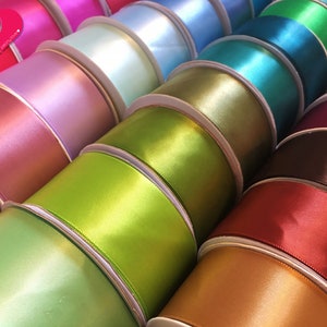 May include: A collection of colorful satin ribbon spools in various shades, including pink, green, blue, and brown. The ribbons have a glossy finish and are neatly rolled on cardboard spools. The image showcases a variety of colors, perfect for crafting and decorative projects.